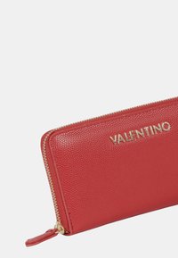 Portafoglio in pelle rossa con finitura texture, logo "VALENTINO" in oro e zip dorata. Design compatto con una linguetta laterale per una facile maneggevolezza.