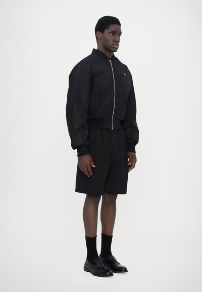 Veste bomber noire avec des poignets côtelés, associée à un short noir cintré. Les accessoires comprennent des chaussettes noires et des mocassins. Texture du tissu lisse.