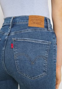 Vaqueros de mezclilla azul con una etiqueta de cuero marrón en la cintura y una pestaña roja en el bolsillo trasero. Presentan un diseño clásico de cinco bolsillos y costuras en forma de corazón.