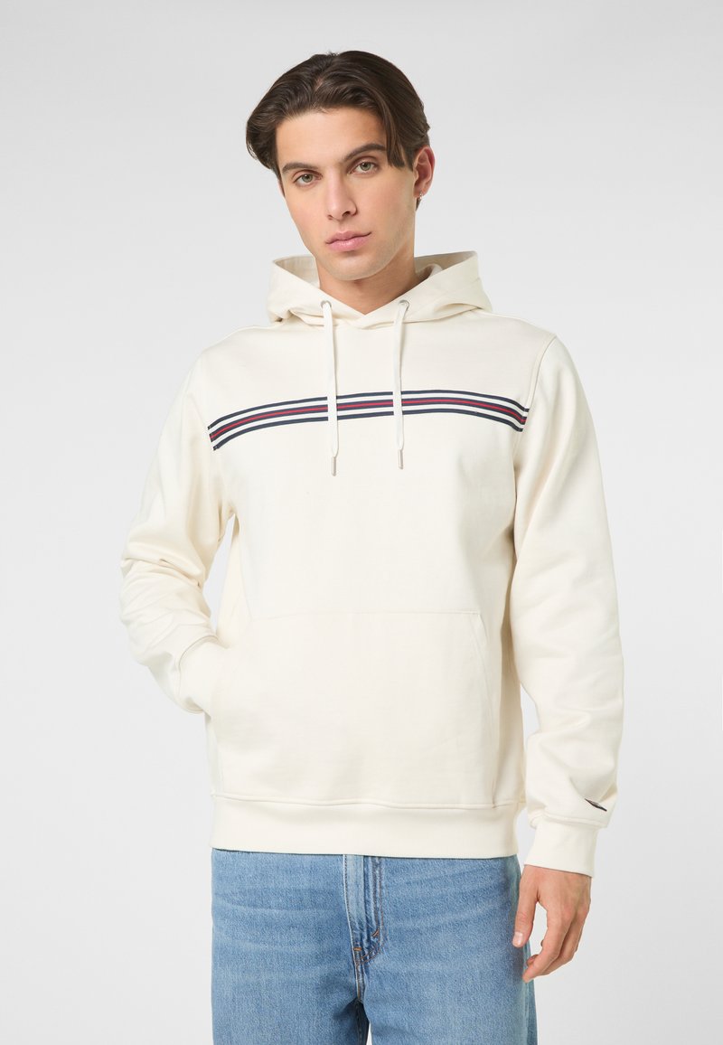 Junger Mann trägt einen cremefarbenen Hoodie mit marineblauen, roten und weißen Streifen quer über der Brust, kombiniert mit hellblauen Jeans, steht mit einer Hand in der Tasche.