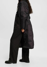 Manteau long matelassé noir à texture brillante, fermeture par boutons, poches latérales et coupe décontractée. Porté avec un pantalon noir à jambes larges et des chaussures robustes.