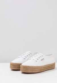 Bílé plátěné espadrilky se silnou spletenou jute podešví. Obsahují kulatou špičku, kovové očko a ploché tkaničky pro bezpečné uchycení.