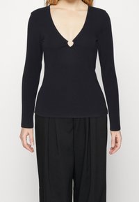 Musta pitkähihaine paita, jossa on syvä V-pääntie ja avainreikä yksityiskohta keskellä etuosaa. Valmistettu sileästä kankaasta, jonka istuvuus on vartalonmyötäinen.