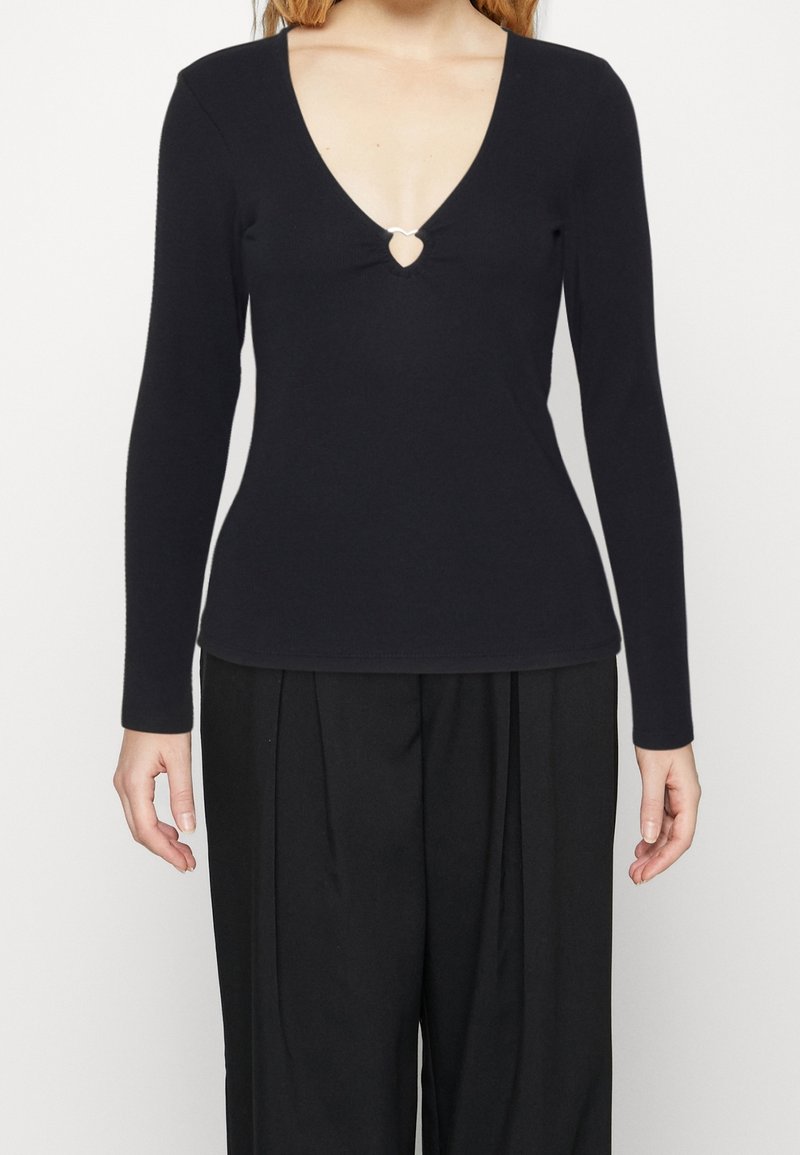 Musta pitkähihaine paita, jossa on syvä V-pääntie ja avainreikä yksityiskohta keskellä etuosaa. Valmistettu sileästä kankaasta, jonka istuvuus on vartalonmyötäinen.