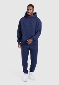 Mann in dunkelblauem Hoodie und passenden Jogginghosen mit weißen Sneakern, der vor einem schlichten hellen Hintergrund steht.