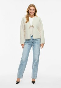 Giacca trapuntata beige con chiusura a cravatta, maniche lunghe e vestibilità ampia. Indossata sopra un top bianco e abbinata a jeans blu e tacchi neri.