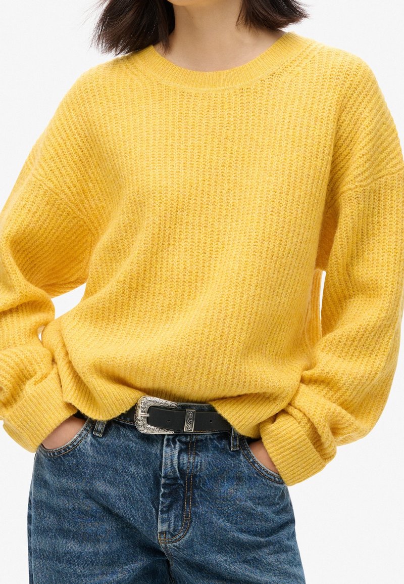 Maglione giallo lavorato a maglia con un motivo testurizzato, scollatura rotonda e maniche oversized, abbinato a jeans blu e una cintura nera.