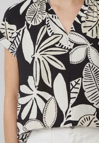 Das kurzärmlige Hemd besteht aus schwarzem Stoff mit großen weißen Blattmotiven, einem V-Ausschnitt und einer strukturierten Oberfläche mit feinen Knitterdetails.