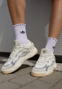 Zapatillas deportivas en crema y plata con tejido texturizado y suela gruesa. Calcetines blancos con bordes rizados y un logo de Adidas.