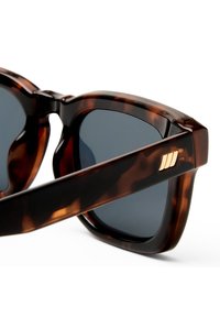 Γυαλιά ηλίου με φαρδιές κορνίζες σε σχέδιο χελώνα και σκουρόχρωμους φακούς, με τρεις χρυσές κάθετες ρίγες στο μπράτσο κοντά στον μεντεσέ.