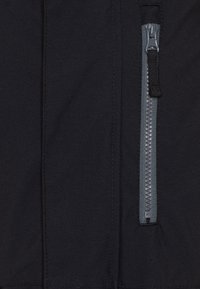 Giacca in tessuto nero con zip in plastica grigia sul lato destro, con cuciture visibili e una texture liscia.