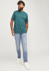 Teal cotton t-shirt with white logo text, paired with light blue denim jeans and white sneakers. Model standing and smiling.