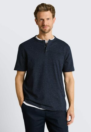 Man met lichtbruin haar en baard, die een donkerblauw henley-shirt met korte mouwen en zwarte broek draagt, staand met één hand in de zak.