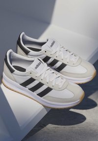 Weiße und beige Adidas-Sneaker mit schwarzen Streifen und Gummisohlen, die auf einer weißen Oberfläche liegen und Schatten auf einen Betonboden werfen.