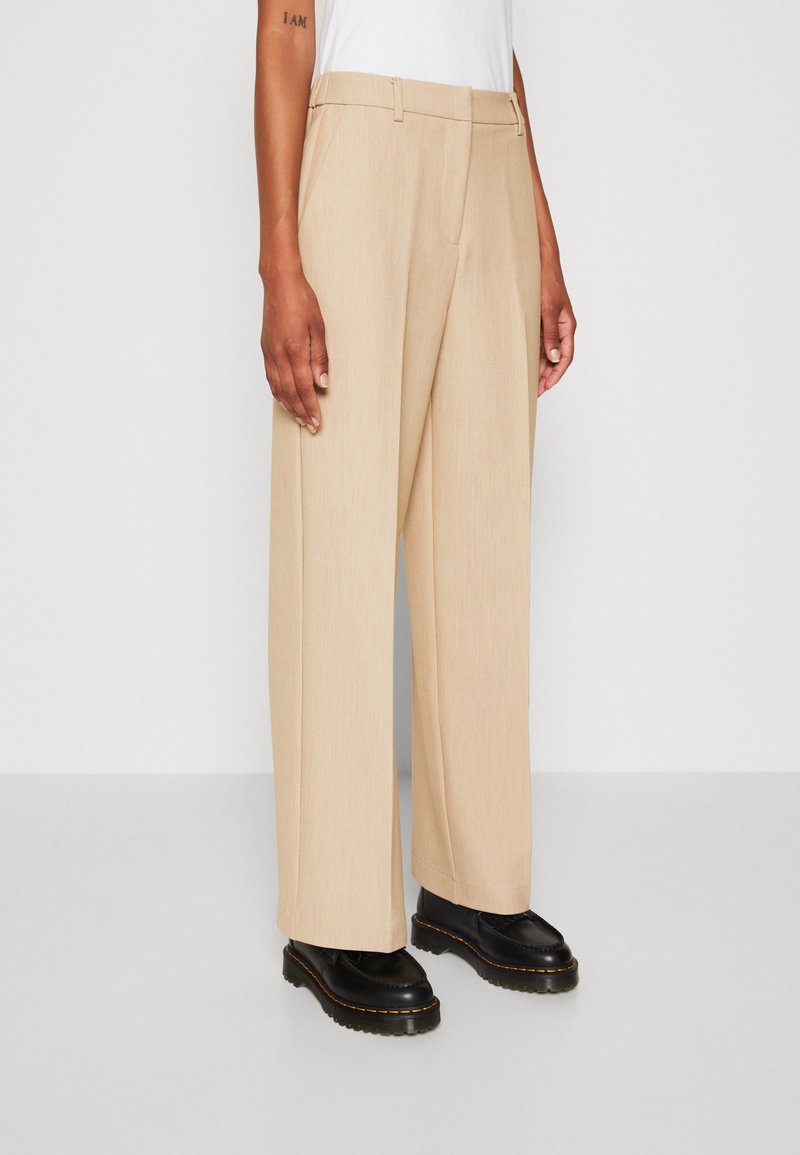 Personne portant un pantalon beige à jambes larges et des chaussures noires épaisses, debout devant un fond blanc uni.