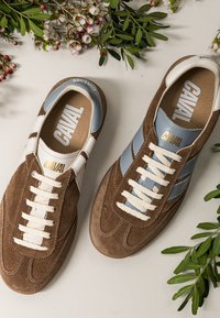Paire de baskets en daim marron et bleu avec des lacets blancs étiquetés "CAVAL", entourée de petites fleurs roses et blanches sur une surface blanche.