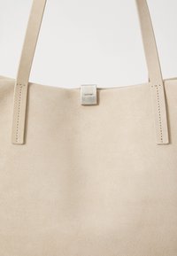 Beige ruskinds tote-tasker med to lange håndtag, prydet med en sølvfarvet metalplade med "Calvin Klein" indgraveret. Glat tekstur, minimalistisk design.