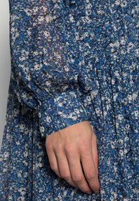 Blaues Blumenlangarmkleid aus leichtem Stoff, mit einem Knopfverschluss am Bündchen und Faltenelement, in einer Mischung aus Blau und Creme.