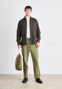 Gestreiftes dunkelbraunes Hemd mit einem weißen T-Shirt darunter, grüne Hose und eine gesteppte olivgrüne Tasche. Schwarze Schuhe runden das Outfit ab.