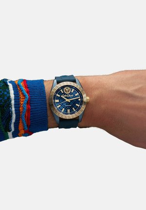 Orologio blu con dettagli dorati, quadrante rotondo, bezel con motivo e cinturino in gomma. Indossato su un polso con un maglione multicolore.