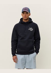 Marinefarbener Sweatshirt mit Kordelzugkapuze, Fronttasche und weißem "LD"-Logo. Kombiniert mit blauen gestreiften Shorts und einer Farbverlaufskappe.