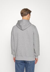 Sudadera gris con capucha, de mangas largas, con puños y bajo acanalados. Fabricada con un tejido suave, sin patrones ni acentos visibles. Vista trasera.