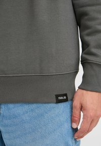 Sudadera gris con puños y dobladillo acanalados, que presenta una etiqueta negra con texto blanco. Primer plano de un tejido suave y texturizado con jeans de mezclilla.