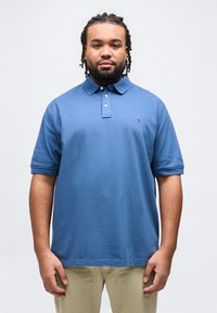 Άνδρας με πλεξούδες στα μαλλιά και γένια, που φοράει μπλε πόλο με κοντά μανίκια και μπεζ παντελόνι, στέκεται μπροστά σε απλό ανοιχτό φόντο.