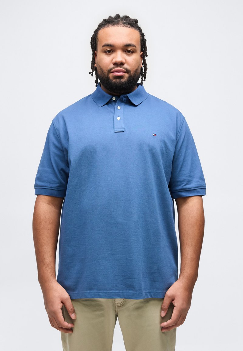 Άνδρας με πλεξούδες στα μαλλιά και γένια, που φοράει μπλε πόλο με κοντά μανίκια και μπεζ παντελόνι, στέκεται μπροστά σε απλό ανοιχτό φόντο.