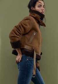 Veste en shearling marron avec un col montant, des poches zippées et une ceinture décorative. Portée avec un jean bleu sur un fond vert.