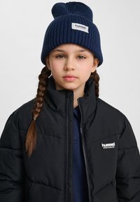 Svart puffjacka med framzipper och logotyp, tillsammans med en marinblå stickad mössa som har en vikbar brätte och etikettdetalj.