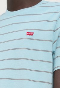 Άνδρας που φοράει ανοιχτό γαλάζιο μπλουζάκι με λεπτές οριζόντιες γκρι ρίγες και κόκκινο πάτς με το λογότυπο Levi's στο στήθος.