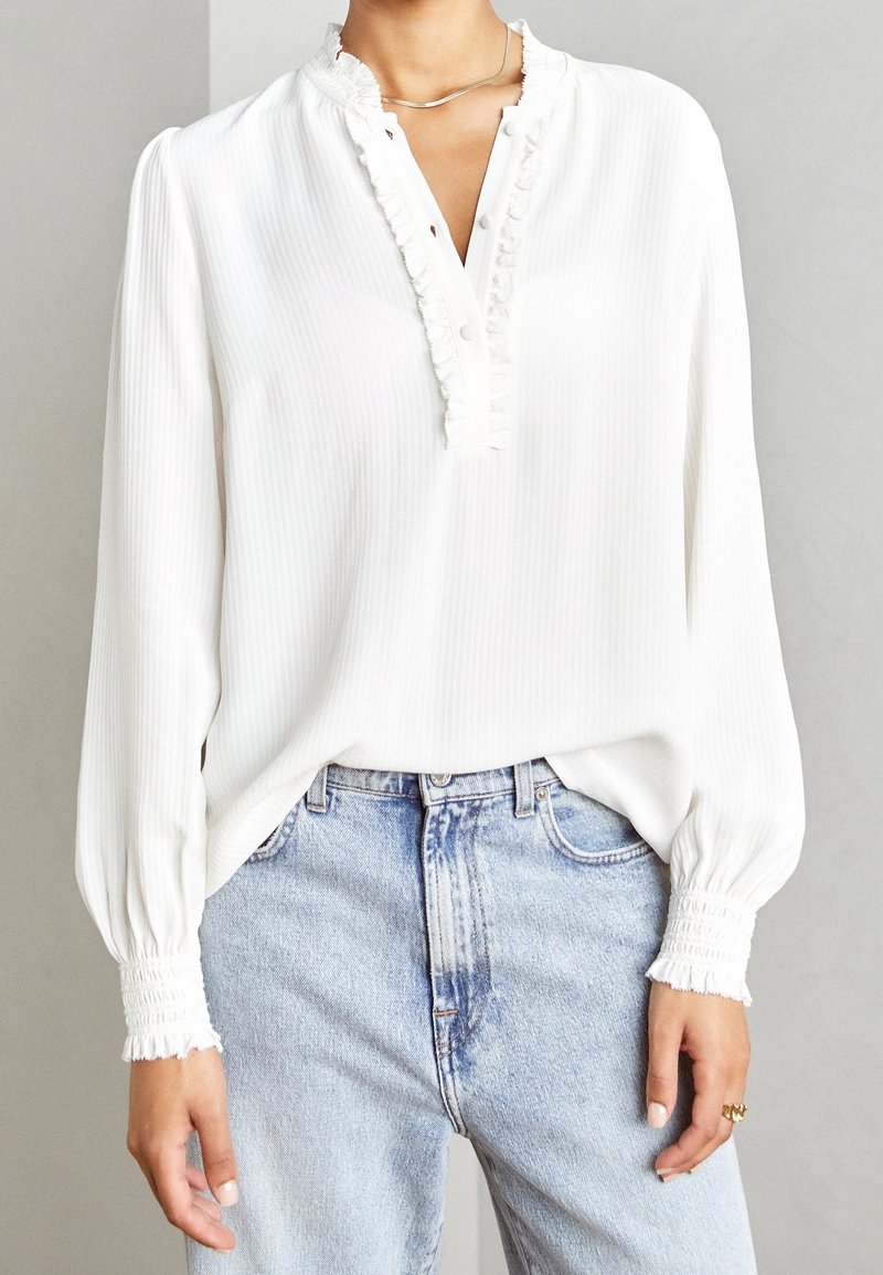 Hvid langærmet bluse med flæsekrave og manchetter parret med lyseblå højtaljede jeans og en simpel sølvhalskæde.