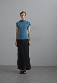 Blå kortermet topp med høy hals, kombinert med et langt svart skjørt. Modell iført svarte sandaler, mot en enkel grå bakgrunn.