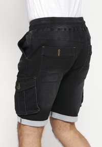 Pantalones cortos de mezclilla negra con una cinturilla elástica, puños enrollados, múltiples bolsillos y detalles de costura amarilla. Adecuados para un estilo casual.