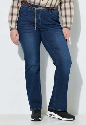 Persoon draagt blauwe jeans met rechte pijpen, een kettingriem, zwart-witte sneakers en een beige geruit overhemd, staande tegen een witte muur.