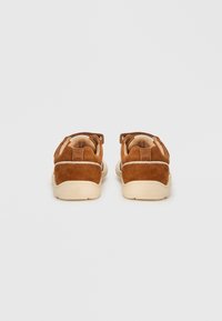 Καφέ πάνινα παπούτσια από σουέντ με μπεζ σόλα, διαθέτοντας επενδυμένο γιακά και απλό, στρογγυλεμένο σχεδιασμό. Η πίσω πλευρά έχει λεπτομέρεια με διακριτικό ραφτό μοτίβο.