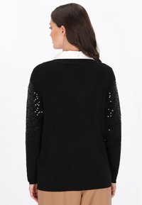 Schwarzer Pullover mit langen Ärmeln und gerippter Textur, der Paillettenakzente an den Schultern aufweist. Kragen unter dem Pullover sichtbar.