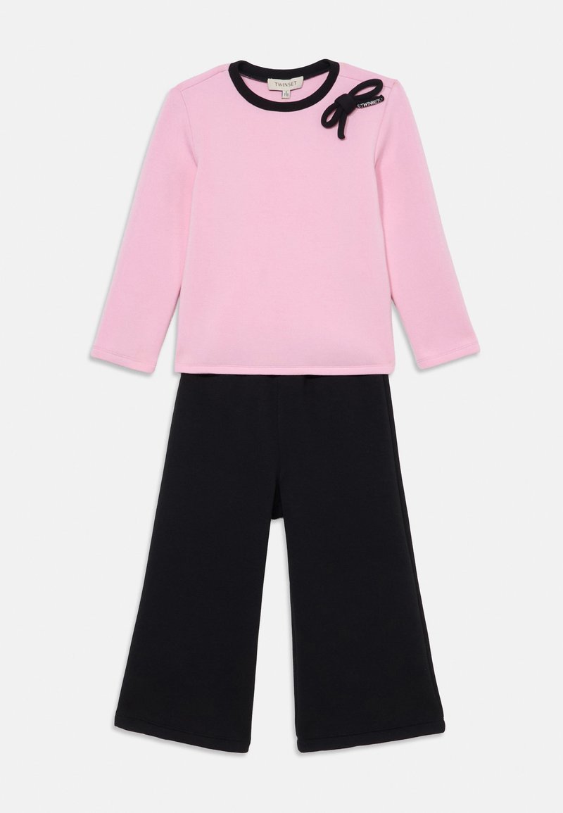 Maglietta rosa a maniche lunghe con rifiniture nere e dettaglio a fiocco, abbinata a pantaloni neri a gamba larga. Entrambi i capi sono realizzati in tessuto morbido.