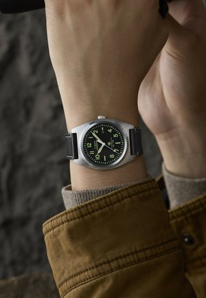 Handgelenk mit einer silbernen Timex Expedition Uhr mit schwarzem Zifferblatt und grünen leuchtenden Zahlen und Zeigern, bekleidet mit einer braunen Jacke und einem beigen Pullover.