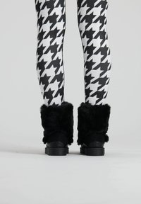 Schwarze, pelzige Stiefel mit einem strukturierten Schaft, kombiniert mit hochkontrastierenden schwarz-weißen Hahnentritt-Leggings. Glatte Sohle und robuste Bauweise.