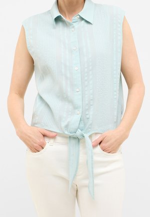 Blouse à manches courtes bleu clair avec rayures verticales et boutons à l'avant, nouée à la taille. Portée avec un pantalon blanc, en tissu doux et texturé.