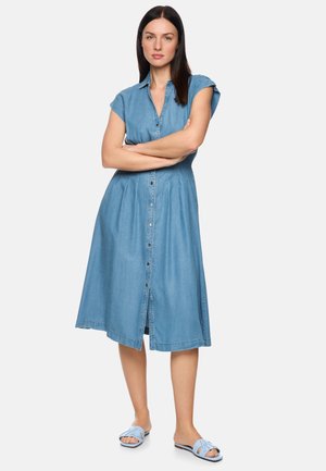 Woman with long dark hair wearing a blue button-up knee-length denim dress and light blue slip-on sandals, standing with arms crossed.