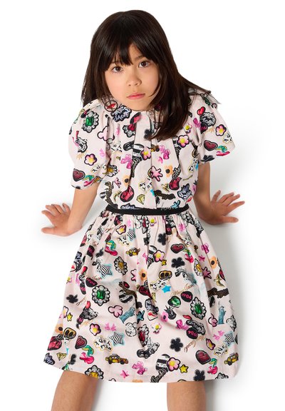 Jeune fille aux longs cheveux foncés portant une robe colorée à motifs variés et ludiques, assise les mains posées sur le sol.