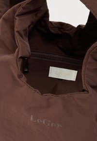 Καφέ υφασμάτινη τσάντα με ορατή τσέπη με φερμουάρ και λευκή ετικέτα στο εσωτερικό που γράφει "LeGer," και το όνομα της μάρκας τυπωμένο στην εξωτερική επιφάνεια.