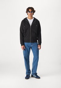 Felpa nera con zip e tasche a canguro sopra una t-shirt bianca, abbinata a jeans blu e sneakers blu con tre strisce.
