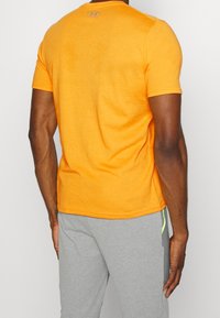 Oranžové tričko s krátkým rukávem vyrobené z měkkého, prodyšného materiálu. Má kulatý výstřih a decentní branding na zadní straně. V kombinaci se šedými kalhotami.