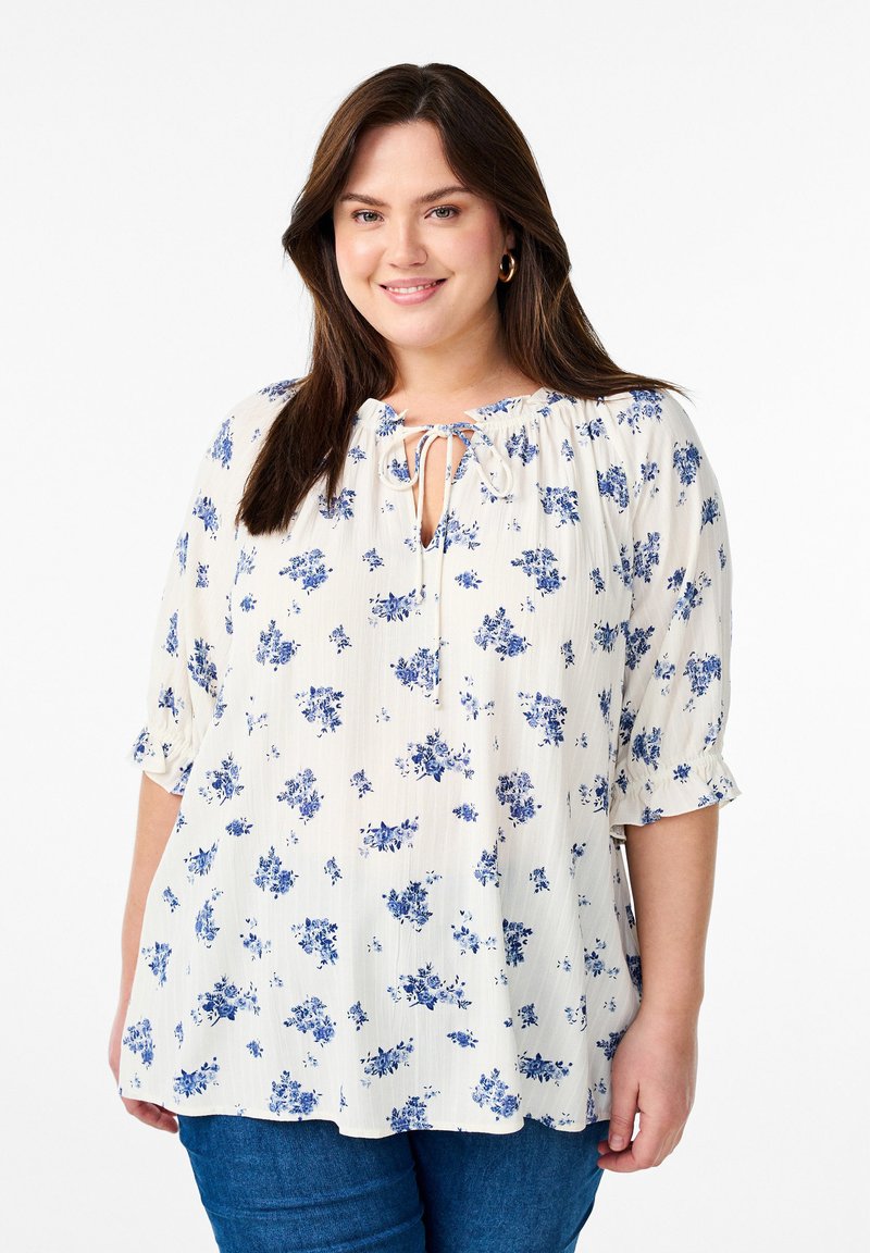 Blusa con motivo floreale color crema e fiori blu, scollatura rotonda con fiocco, maniche 3/4 con polsi elasticizzati, vestibilità comoda, tessuto leggero.