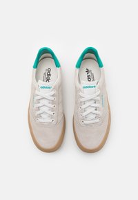 White suede sneakers with gum rubber soles, turquoise accents, and white laces. Features branded insole and logo detailing.
