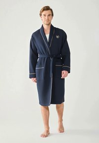 Navy-blaue Bademantel mit Waffelstruktur, kontrastierendem hellblauen Piping, Bindegürtel, zwei vorderen Taschen und einem gestickten Emblem auf der Brust.